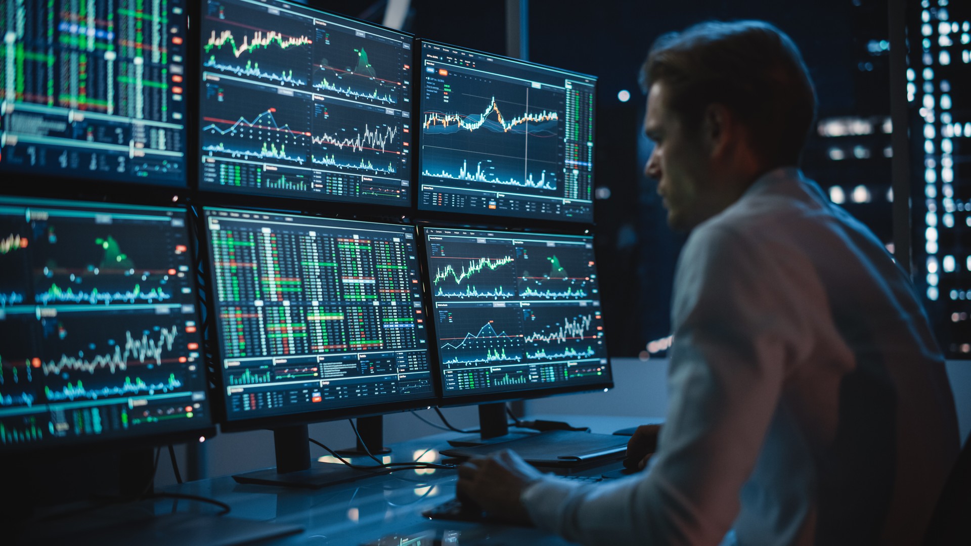 Financial Analyst Working on a Computer with Multi-Monitor Workstation with Real-Time Stocks, Commodities and Exchange Market Charts. Businessman Works in Investment Bank Downtown Office at Night.